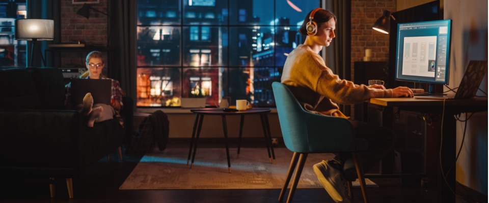 A person sits at a desk with headphones, reviewing documents on a desktop computer, while another individual relaxes on a nearby couch, visually representing the process of researching how to cancel renters insurance and exploring alternative coverage options.