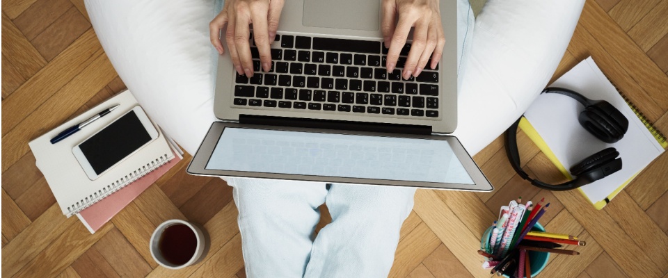 Overhead view of a person sitting cross-legged on a parquet floor, working on a laptop with notebooks, smartphone, headphones, pens, and a cup of coffee nearby, illustrating the comfort of an apartment for remote workers.
