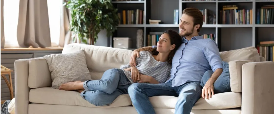 A couple relaxing together on a modern light-colored sofa in a spacious living room, surrounded by bookshelves and natural light, illustrating how flexible rent payment options help renters reduce stress about monthly costs.