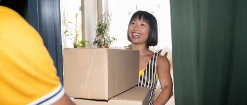 A smiling woman receives two cardboard boxes at her front door from a delivery person, having more flexibility over her budget due to choosing a flexible rent payment option.
