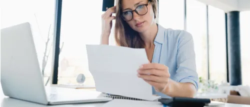 A focused woman with glasses looking at a file and having a laptop on a white table, while researching how to cancel renters insurance.