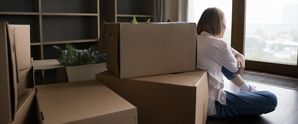 Woman sitting alone by window surrounded by moving boxes, looking out quietly after moving to a new state alone.