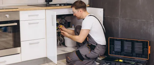 A maintenance worker in overalls uses tools to fix plumbing under a kitchen sink in a modern apartment, handling an emergency repair.