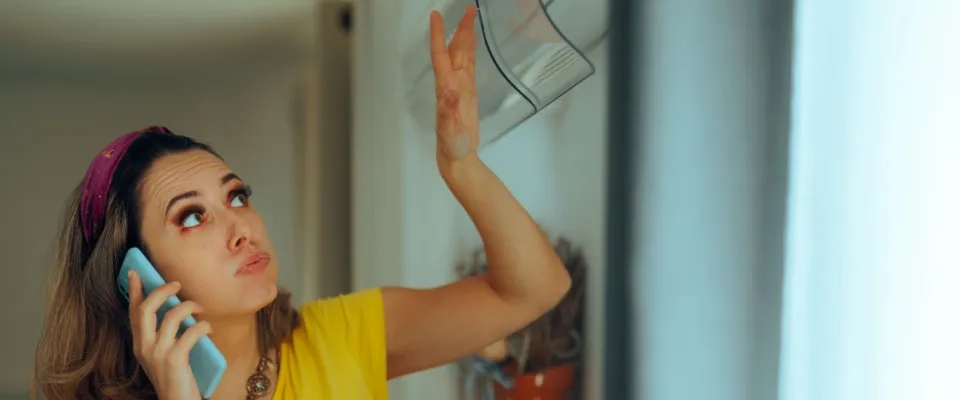 A woman on the phone raises her hand in frustration under a malfunctioning air conditioning unit in her apartment, which requires emergency repairs.