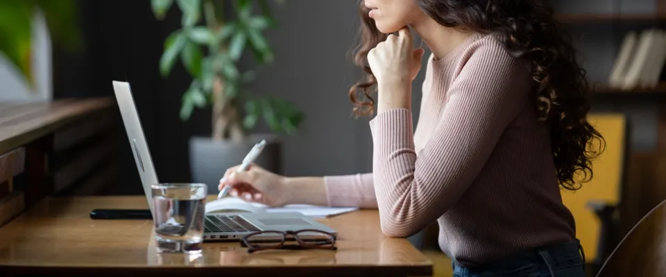 A woman sitting at a wooden desk with a laptop and a notebook, documenting an issue happening in her apartment in need of emergency repairs.