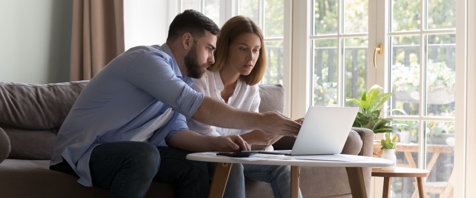 A couple sits together on a sofa in a bright living room, reviewing documents for rent reporting and using a laptop on a coffee table