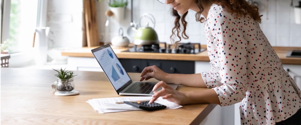 A woman calculates utility bills with a calculator while reviewing charts of her credit score on her laptop in a bright kitchen.