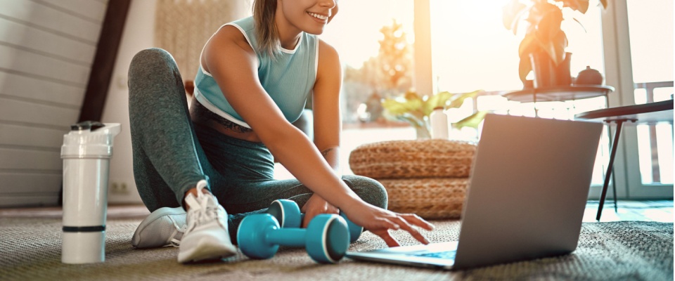 A woman in athletic wear smiles while taking an online fitness class from home, sitting on a sunlit carpet with exercise weights and a water bottle nearby, working out in the morning as an energy-saving tip during summer.