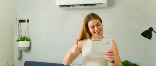 A woman standing in a bright living room holds up an electricity bill and gives a thumbs up beneath a wall-mounted air conditioner, after following energy-saving tips to keep her apartment cool in summer.