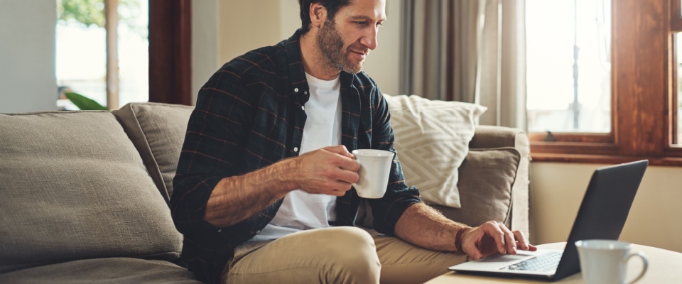A man working remotely on his laptop from the sofa with a drink, using technology for blogging, company reviews, or small business tasks at home, reflecting modern lifestyles and the search for a roommate.