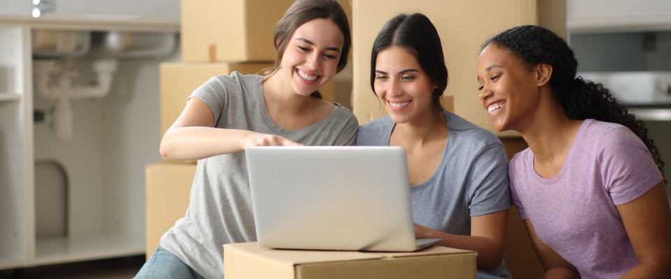 Three happy roommates checking a laptop together while moving into a new place, capturing the excitement and teamwork of the search for a roommate.