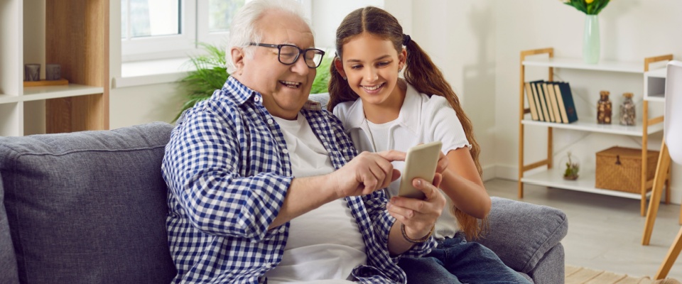 A touching moment between grandfather and granddaughter as they share smiles over a smartphone — a beautiful example of connection and bonding in a multi-generational household through modern technology.