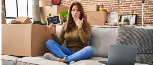 Woman surrounded by moving boxes holding a “New Home” sign, representing apartment hunting mistakes renters should avoid during peak rental season.