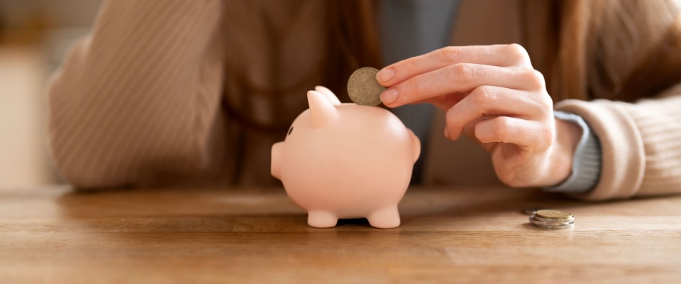 A person saving money by putting coins into a pink piggy bank on a wooden table in a cozy indoor setting, reflecting smart budgeting and financial planning during the search for a roommate.