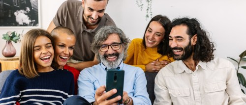 A multi-generational family sits closely on the sofa at home, sharing a moment around a mobile phone — showcasing connection, technology use, and everyday life in a multi-generational household.