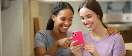 Two happy roommates checking a phone together in the kitchen while moving into their new home, illustrating the search for a roommate.