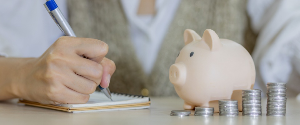 A woman placing a coin into a piggy bank alongside neatly stacked coins symbolizes smart budgeting and saving—an important habit when moving near a college town, where managing expenses and planning ahead can make student life more affordable and stress-free.