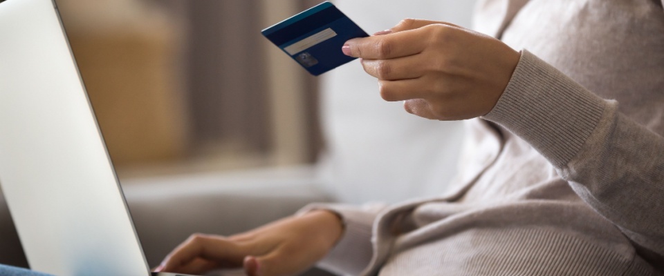 Close-up of a young woman sitting on a sofa at home, applying online for rentals with a laptop and credit card in hand, carefully managing expenses while factoring in an application fee.