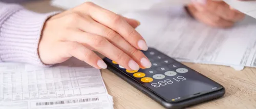 Close-up of a woman’s hand using a calculator app on a smartphone with utility bills on the table, illustrating the process of comparing electricity plans to find the most cost-effective option.