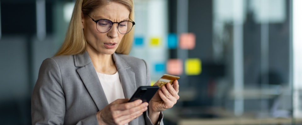 Businesswoman in a gray suit holding a smartphone and credit card in a modern office, suspecting the possibility of identity theft and bank fraud.