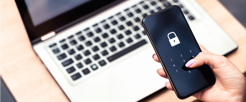 Person entering a passcode on a smartphone with a lock icon displayed, sitting at a desk with a laptop, illustrating steps to prevent identity theft through device security.