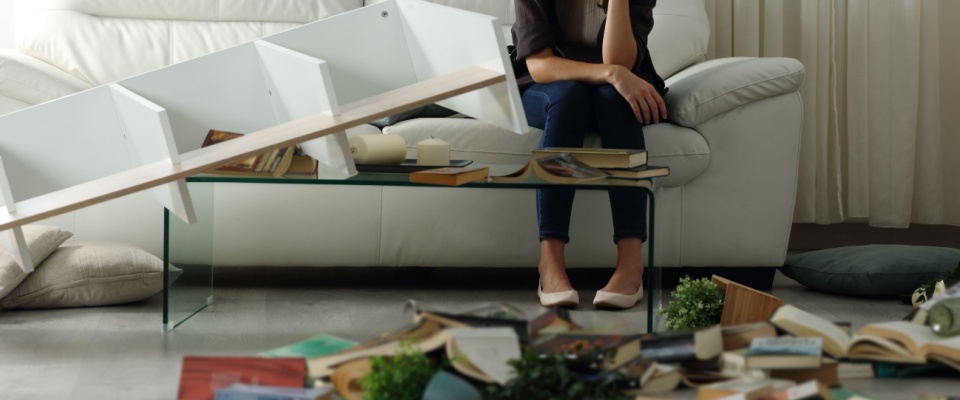 A woman sits on a white sofa in a disrupted living room with fallen shelves, scattered books, and damaged decor, visually illustrating the need for renters insurance coverage to protect high-value items from unexpected accidents or disasters.