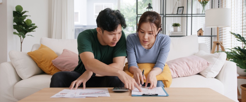 Couple sitting on a sofa in a bright living room, reviewing utility bills and documents with a calculator on the coffee table, demonstrating the importance of comparing electricity plans.