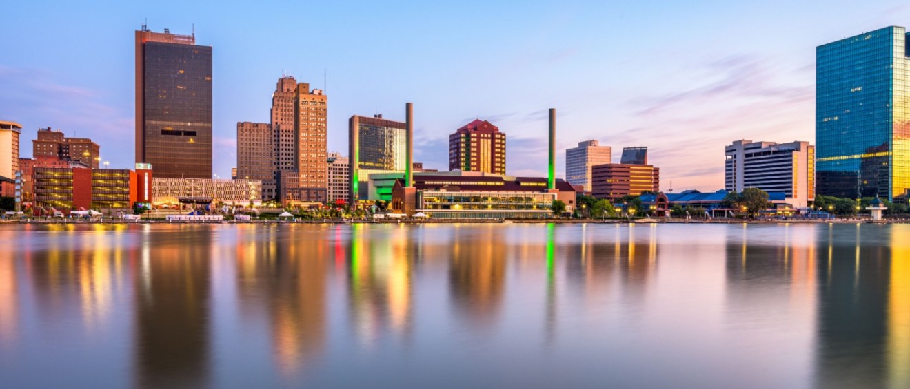 Toledo, OH downtown skyline reflecting on the Maumee River at dusk — a growing city where many people explore affordable neighborhoods for a balance of community and cost of living.