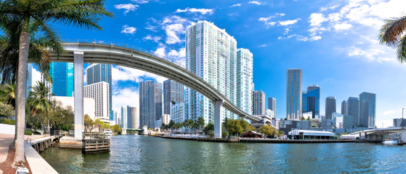 Panoramic view of Miami’s downtown skyline with the elevated Metromover track crossing the Miami River, highlighting public transit options near high-rise apartments.