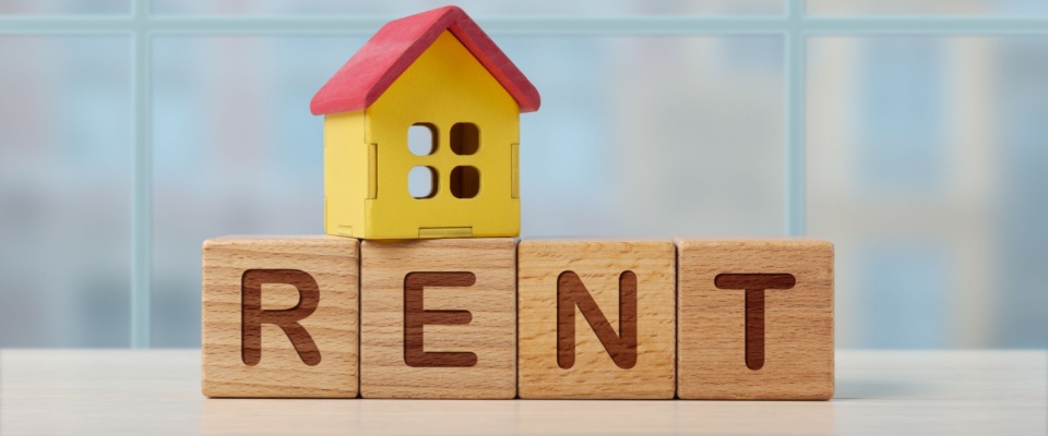 A model house on wooden blocks spelling “RENT” with a red roof and yellow walls. It highlights rental housing and the importance of the lease commencement date, which marks the official start of a rental agreement.