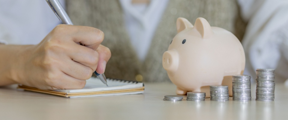 A woman’s hand places a coin into a piggy bank beside a stack of coins. This image reflects saving habits tied to rent, mortgages, and the lease commencement date, which marks the beginning of financial responsibility for a new home.