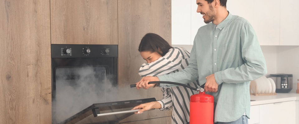 Man using a fire extinguisher while smoke rises from an oven and woman covers her face, showing the importance of fire prevention and kitchen safety.