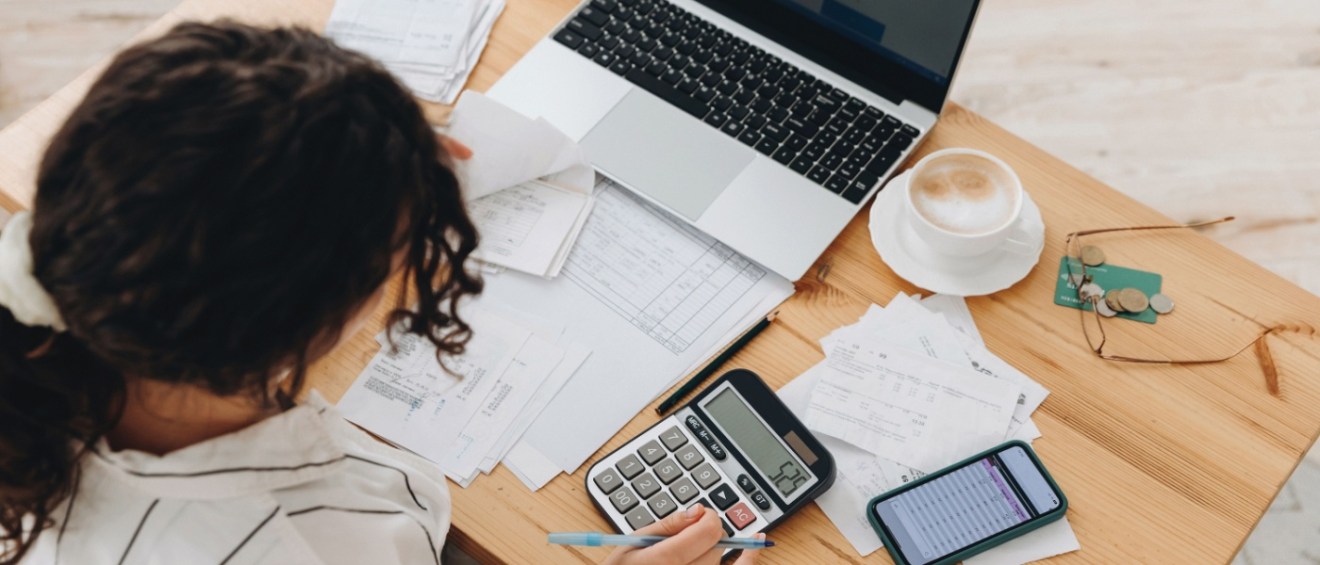 Woman using a calculator and laptop to review multiple invoices and payments, analyzing household expenses for utility billing.