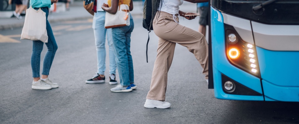 A group of passengers waiting and chatting happily at a bus stop, with a woman boarding the bus — a lively scene that captures the everyday energy of Baltimore public transit.