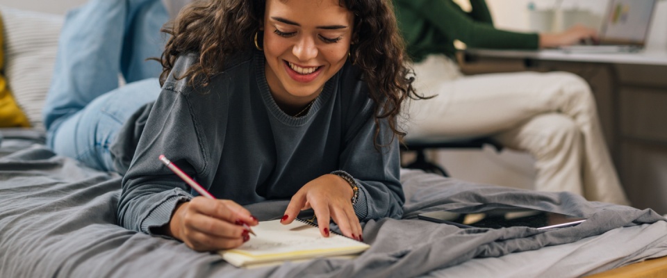 Smiling teenage girl doing homework in her dorm room, highlighting independence and student life, suitable for renting a dorm or apartment without a landlord reference.