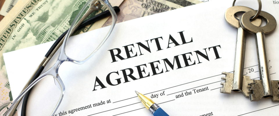  Alt text for the image: A close-up view of a rental agreement document with the title "RENTAL AGREEMENT" prominently displayed, resting on a surface with scattered dollar bills, a pair of reading glasses, a pen, and a set of keys, illustrating the financial and legal aspects of renting with a cosigner.