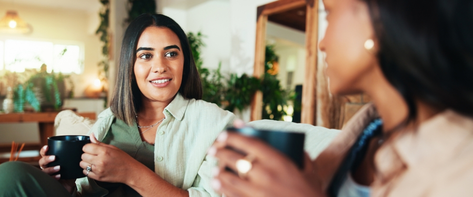 Two women, each holding a dark mug, engage in conversation in a sunlit living room with abundant plants, with the woman on the left in clear focus, embodying a serious discussion about who can be a cosigner.