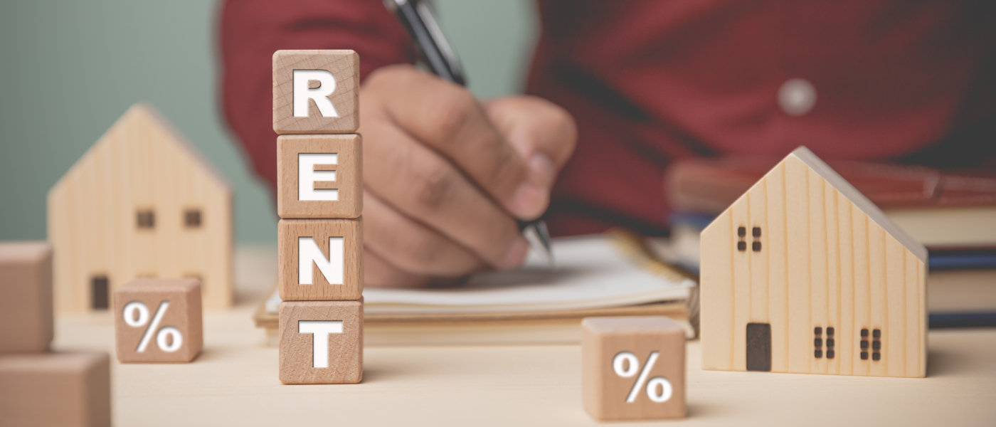 Close-up of wooden blocks spelling “RENT” alongside miniature house models and percentage symbols, with a person writing in the background, representing the process of negotiating rent concessions and understanding rental terms.