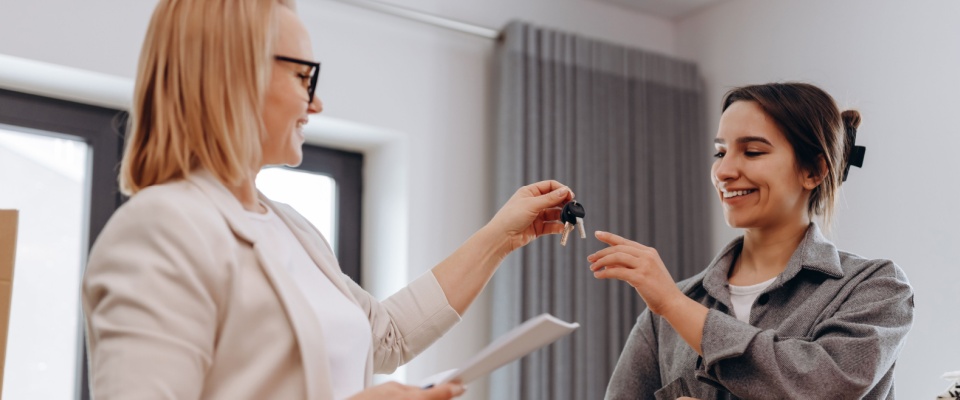 Close-up of female real estate agent handing over keys to a happy young woman, symbolizing the milestone of becoming a first-time renter.