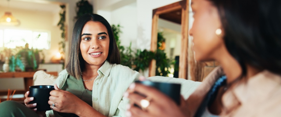 Friends sitting on a sofa in a cozy apartment, drinking coffee and talking together in the living room, representing renter resolutions around social connection, balance, and comfortable home living.