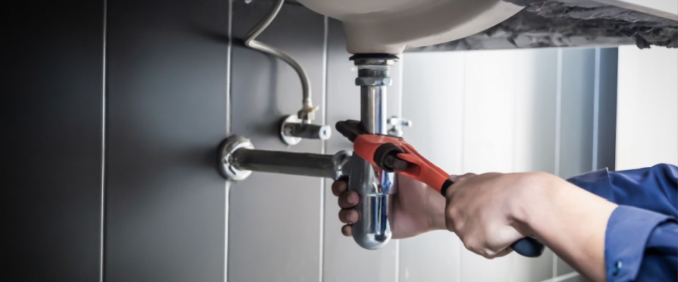 Apartment tour maintenance scene: Plumber using a wrench to repair a water pipe under the sink, fixing a bathroom leak with a red-handled tool.