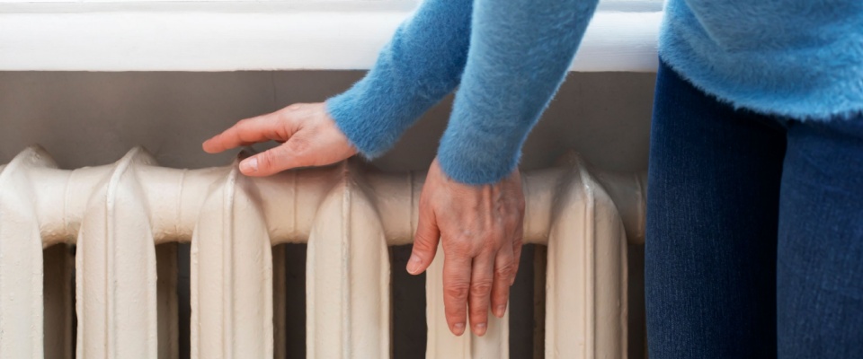 Apartment tour scene showing a woman warming her hands near a hot water radiator in a cozy interior.