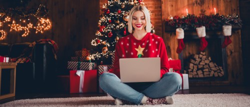 Woman sitting on the floor in a festive holiday living room, using her laptop to organize her next apartment search in front of a decorated Christmas tree and wrapped gifts.