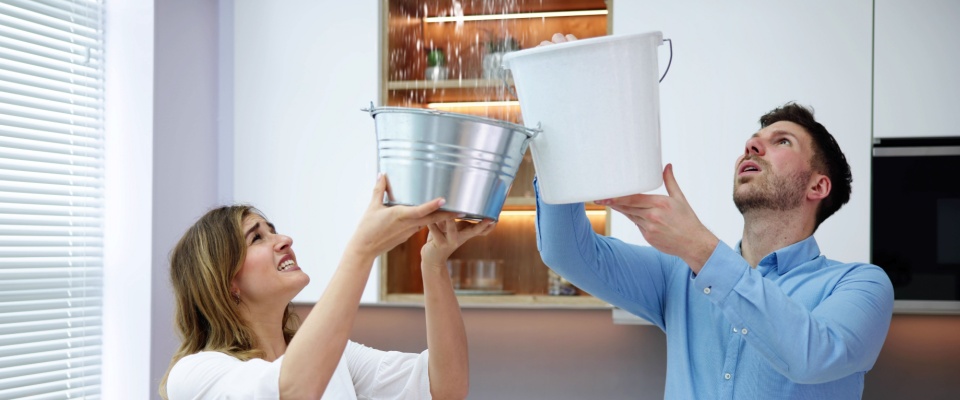Couple holding buckets under a leaking ceiling to catch water, showing situations where renters insurance coverage add-ons may help cover damage