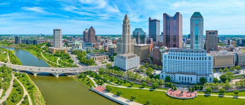 New apartments in Columbus showcased in an aerial panoramic view of Columbus, Ohio, featuring a bridge over the Scioto River beneath a bright blue sky with scattered clouds.