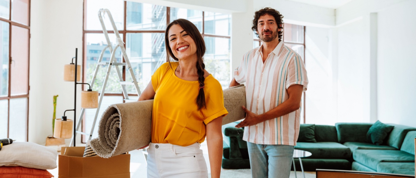 Couple holding a rolled rug, moving into a new apartment with boxes and furniture, highlighting renter essentials and the importance of renters insurance for protecting belongings during a move.