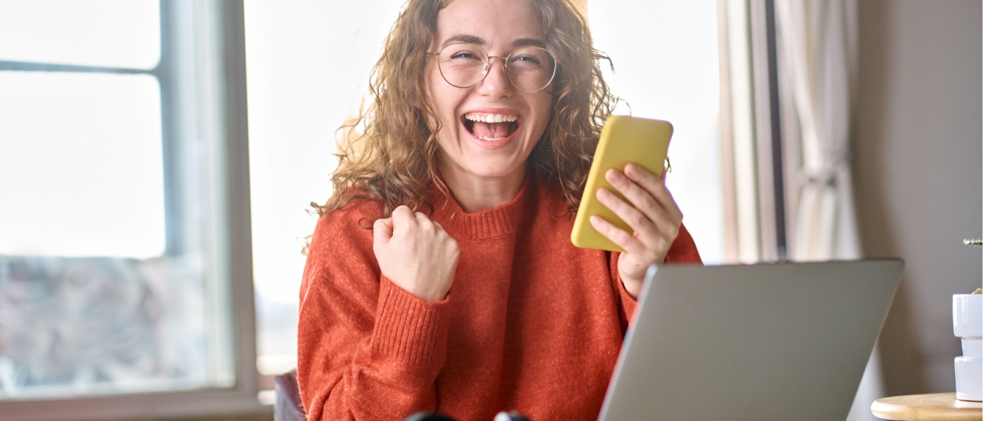 Smiling renter celebrating on her phone while using a laptop, representing earning points and benefits through Rentcafe Rewards for renter essentials and everyday expenses.
