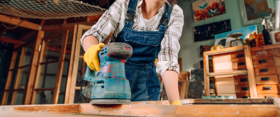 Person sanding a wooden surface in a workshop as part of creating upcycled furniture from reclaimed materials.