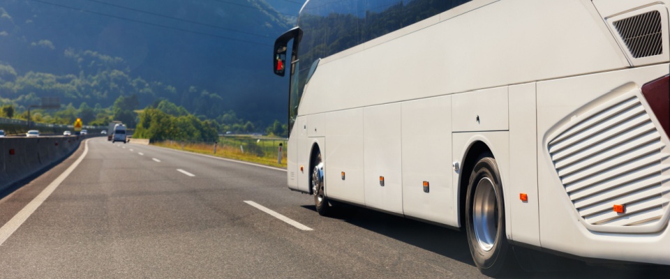 Luxury tour bus driving along a scenic highway with mountain views on a sunny day, representing long-distance travel and public transit options.