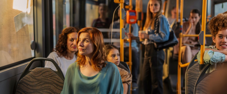Group of passengers sitting and standing on a city bus during a sunny commute, illustrating everyday public transit in an urban area.